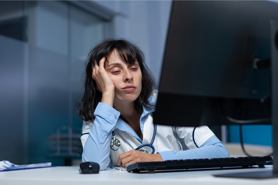 Tired clinician reviewing EMR documentation and charting at a computer after hours, illustrating the administrative burden in healthcare and how AI medical scribe tools like ScribePT help reduce manual charting time.