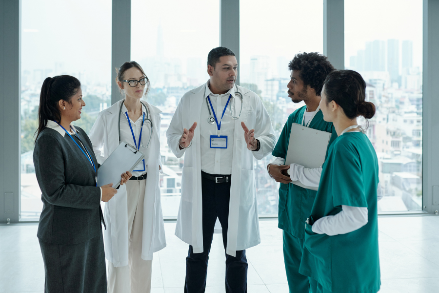 A diverse group of five healthcare professionals and administrators in a modern city office discussing EMR AI adoption strategies and clinical documentation challenges. The team—including a doctor in a lab coat gesturing to colleagues in scrubs and administrative blazers—evaluates the strategic choice between building a complex in-house AI solution versus a rapid integration via ScribePT’s white-label and API options. They are focused on overcoming clinician resistance and documentation debt by choosing ScribePT’s ambient, specialty-specific AI to streamline workflows and improve EMR retention.