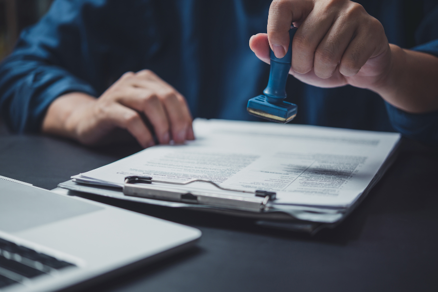 A professional close-up of a hand applying a blue rubber stamp to a document, symbolizing the rigorous verification and independent third-party validation required for a SOC 2 Type II attestation. This visual represents the transition from a "Type I" design review to the "Type II" operational proof required by EMR vendors and healthcare technology partners to ensure long-term data security and HIPAA compliance.