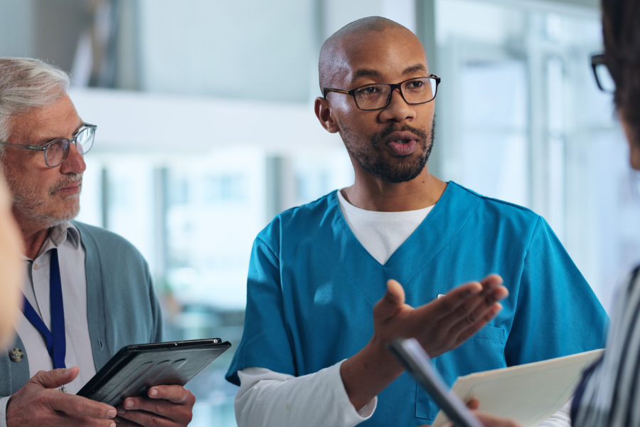 An EMR engineering lead stands with an older product consultant and a clinical partner in a brightly lit medical software office, detailing the low technical lift and rapid deployment advantages of integrating ambient AI documentation APIs to transform raw clinical conversation into structured medical notes.