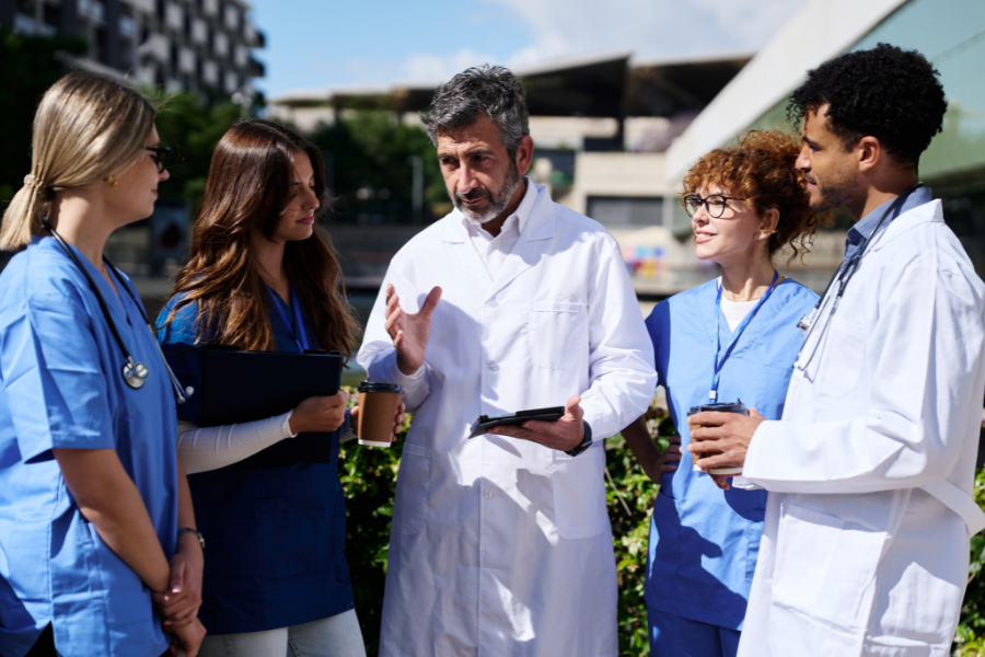 A group of healthcare providers and clinic leaders in white coats and scrubs holding coffee cups while discussing workflow efficiencies, illustrating how practices adopt ScribePT’s secure ambient AI scribe to achieve rapid payback and positive ROI within 30 days.