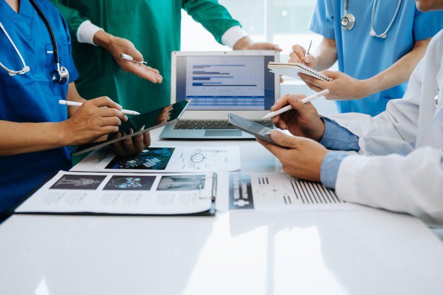 A multidisciplinary team of medical clinicians and healthcare IT consultants collaborating over clinical documentation, digital data charts on a laptop, and tablets to evaluate how to scale AI in healthcare documentation from pilot to enterprise impact with a secure, integrated ambient AI scribe solution like ScribePT.
